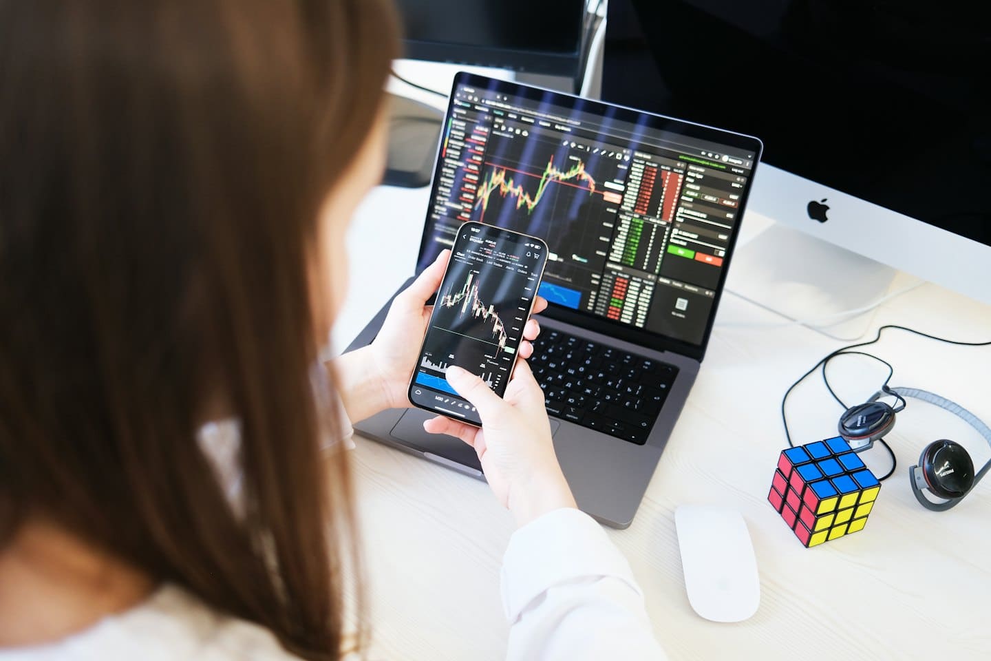 Person trades stocks on phone and laptop in office.